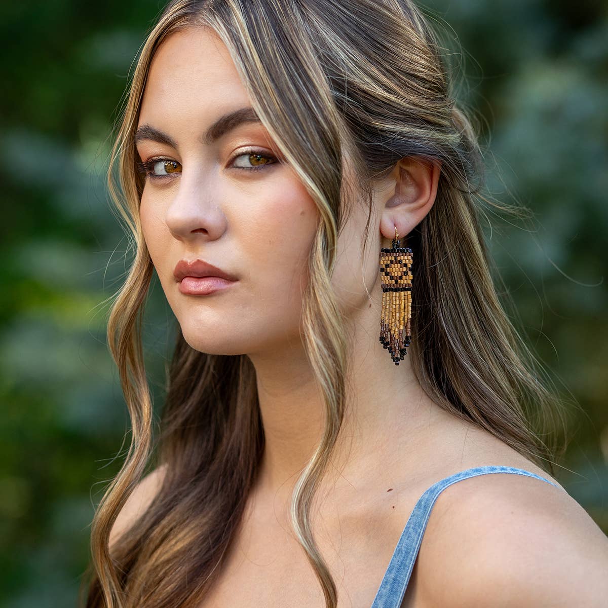 Woman wearing a pair of gold tassel earrings with a blurred green background