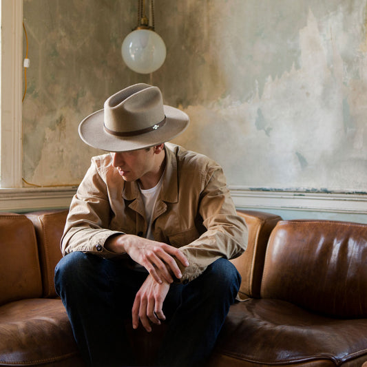 Man wearing a beige hat and coat sitting on a brown leather couch in a room with vintage decor.